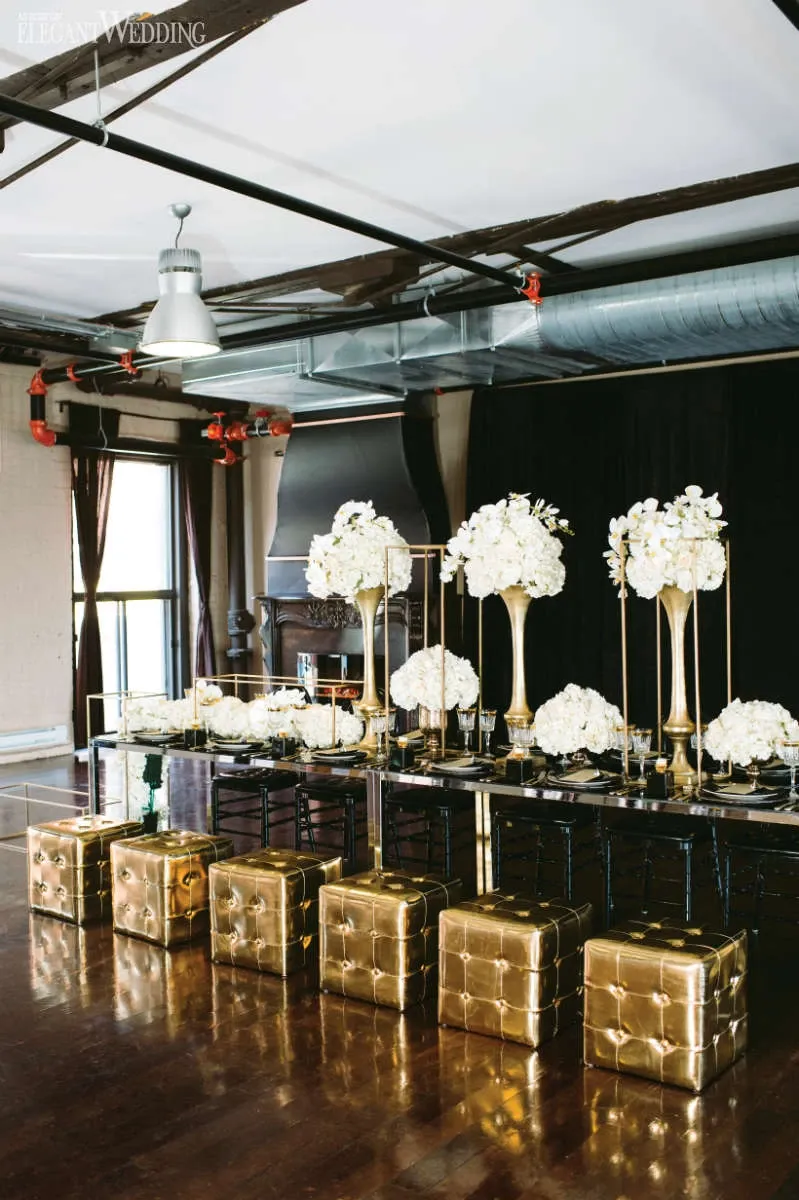 The image showcases a sophisticated wedding floral arrangement by La Grâce des Fleurs, a renowned wedding florist categorized under Wedding Florists on Mariage Elegant, Canada. The setting features a beautifully decorated reception area with elegant, tall gold vases filled with lush white flowers, including roses and hydrangeas, arranged atop a long, polished black table. Gold cube seats are lined neatly in front, enhancing the luxurious theme. The backdrop includes industrial-style elements with exposed beams and a dark curtain, creating a chic and modern wedding ambiance.