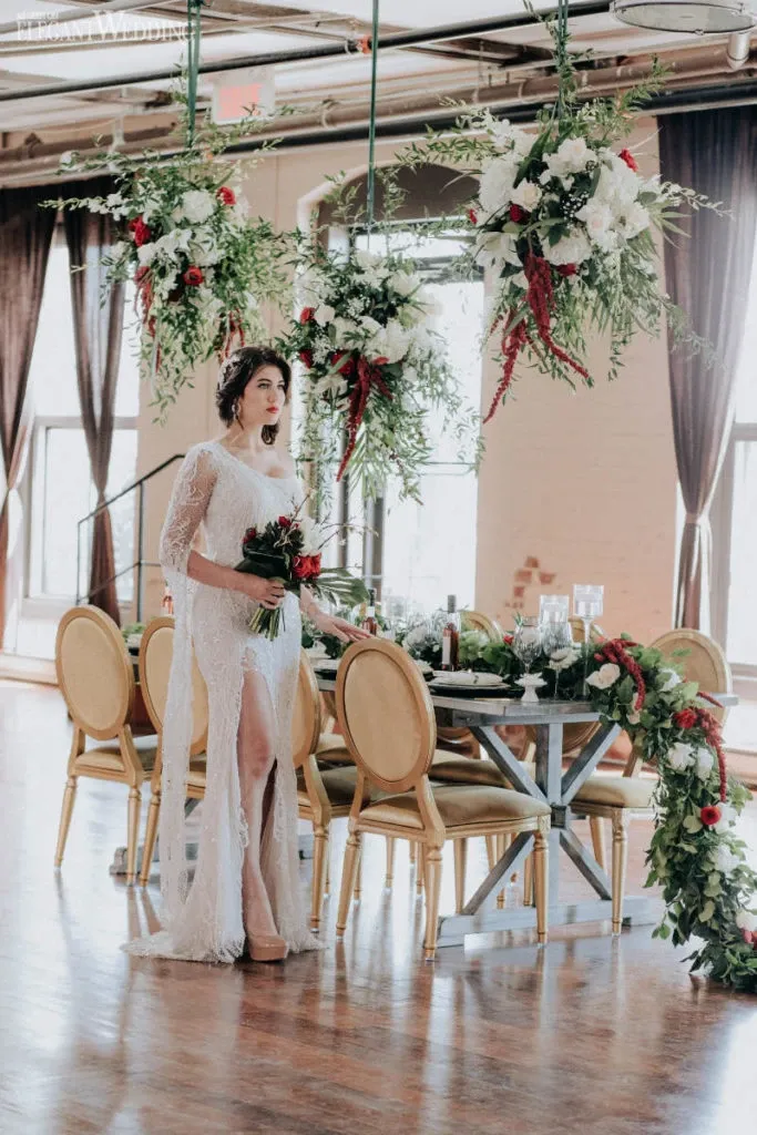 In an elegantly decorated wedding setting by La Grâce des Fleurs, a wedding florist, a bride stands beside a rustic wooden table adorned with a lush floral arrangement. The bride is wearing a white lace wedding gown with intricate designs, holding a bouquet of red and white flowers. Above her, hanging floral displays featuring greenery and red accents complement the arrangements on the table, enhancing the romantic ambiance. The venue features large windows and soft, natural lighting, providing a warm and inviting atmosphere for the celebration.