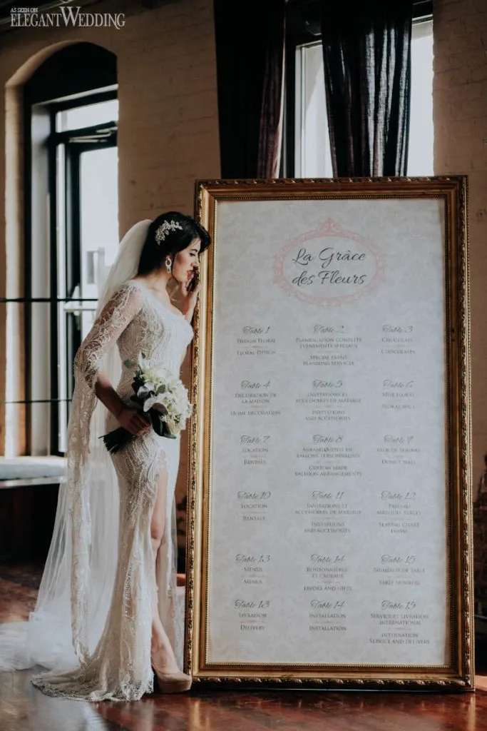 In this elegant image for Mariage Elegant, Canada, a bride in a stunning, lacy white gown and veil stands beside an ornate gold-framed floral arrangement sign by La Grâce des Fleurs, highlighting their expertise in the wedding florists category. She holds a bouquet of white flowers, perfectly complementing the sophisticated scene. The sign displays a detailed seating plan, adding a sophisticated and organized touch that reflects the professional services offered by this vendor for weddings.