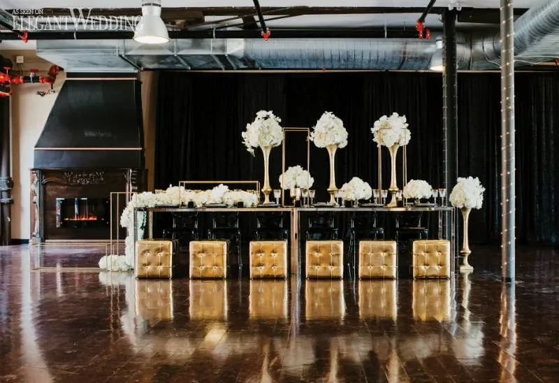 The image features an elegantly decorated wedding venue styled by La Grâce des Fleurs, a wedding florist listed in the Mariage Elegant vendor directory in Canada. The central focus is a luxurious banquet table adorned with tall, gold flower stands overflowing with white blooms. The table is set against a dark backdrop, enhancing the vibrancy of the white flowers. Golden, tufted stools are neatly arranged along the table, adding a lavish touch to the setup. In the background, there is a modern fireplace, contributing to the sophisticated ambiance of the setting.