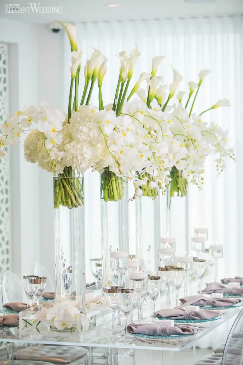 In this elegant image showcasing the work of La Grâce des Fleurs, a distinguished vendor in the Wedding Florists category, tall floral centerpieces are arranged on a beautifully decorated table. The centerpieces feature white calla lilies and hydrangeas, complemented by delicate orchids, all placed in clear vases that add a sense of height and grace to the setting. The table is set with polished glassware and pastel-colored napkins, evoking a sophisticated and romantic ambiance. This arrangement perfectly embodies the luxurious and elegant style that Mariage Elegant in Canada is known for.