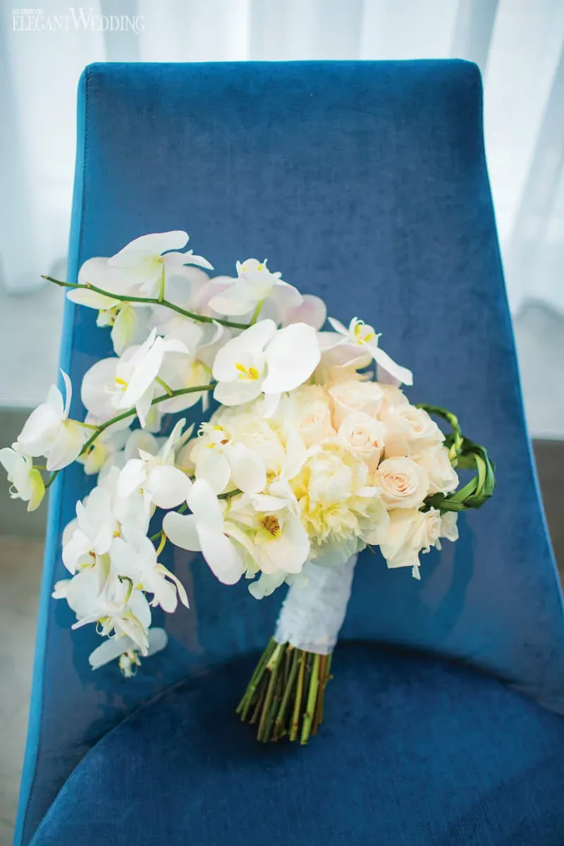 A beautiful bridal bouquet by La Grâce des Fleurs, a distinguished name in wedding florists, is elegantly displayed on a rich blue chair. The arrangement features cascading white orchids paired with soft peach and cream roses, creating an exquisite and romantic floral masterpiece. The delicate wrapping adds an elegant touch, making it an ideal choice for a sophisticated wedding celebration.
