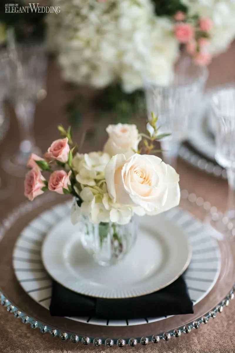 A beautifully arranged floral centerpiece by La Grâce des Fleurs, a renowned wedding florist, is elegantly displayed on a wedding reception table. The arrangement includes soft blush pink roses, white roses, and delicate greenery set in a clear glass vase. The centerpiece sits atop a fine china plate with a black napkin, surrounded by crystal stemware and elegant glass charger plates with beaded rims. In the background, white hydrangeas and more pink roses further enhance the romantic and sophisticated ambiance. This exquisite floral design exemplifies the artistry and attention to detail provided by La Grâce des Fleurs for weddings.