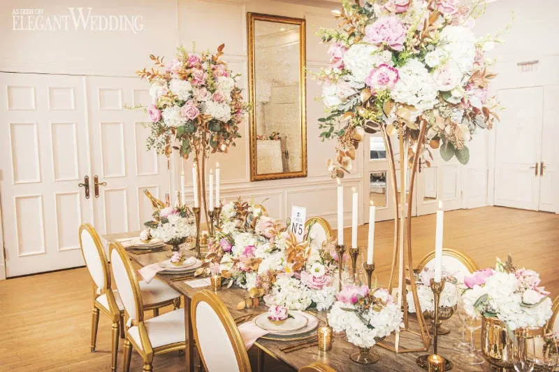 The image showcases an elegantly arranged wedding dining setup by La Grâce des Fleurs, categorized under Wedding Florists. The table is adorned with tall, opulent floral centerpieces featuring a mix of pink, white, and peach blooms, accented with greenery. Gold-rimmed chairs surround the wooden table, which is neatly set with matching gold flatware and white plates. Slim, white taper candles in gold holders add a touch of sophistication. The backdrop features cream-colored walls and large mirrors, creating a spacious and luxurious ambiance, perfect for a wedding setting.
