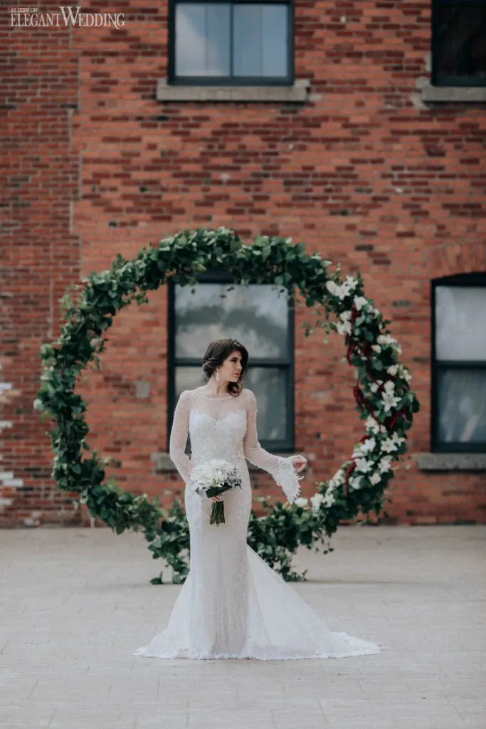 A bride in a lace wedding dress stands in front of a large circular floral arch, elegantly designed by La Grâce des Fleurs, a wedding florist. The arch is adorned with lush greenery and white flowers, complementing the rustic brick building in the background. This setting by La Grâce des Fleurs, a featured vendor in the wedding florists category, showcases an exquisite blend of elegance and natural beauty, ideal for a wedding ceremony backdrop.