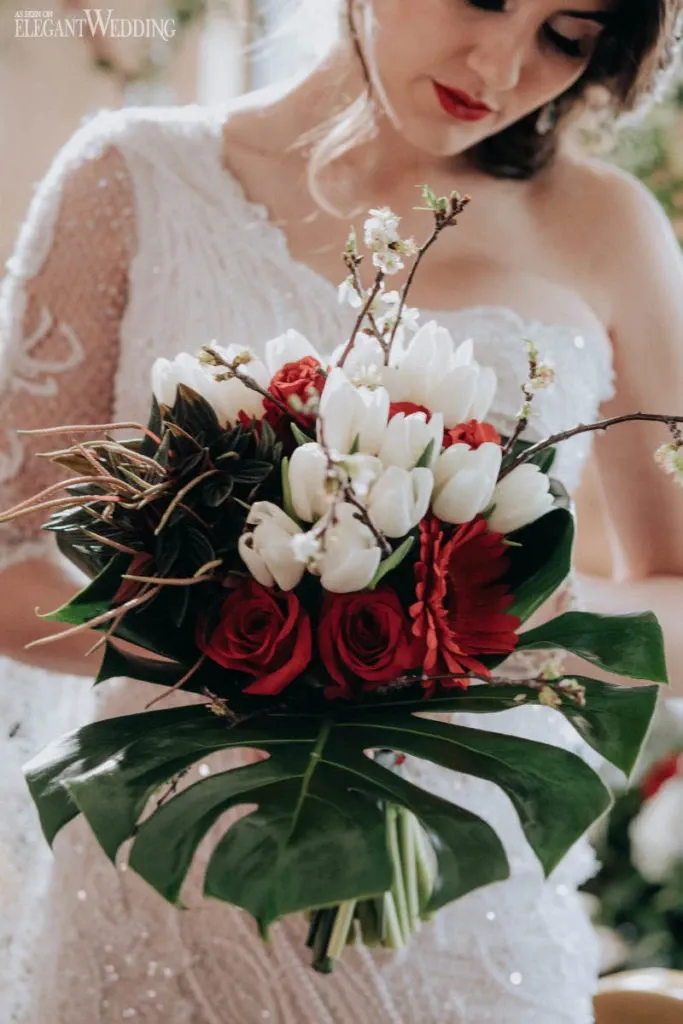 A bride holds a beautifully arranged bouquet by La Grâce des Fleurs, a vendor in the Wedding Florists category. The bouquet features a mix of white tulips, vibrant red roses, and deep red gerbera daisies, accented with green foliage and delicate branches. The bride is dressed in a white lace wedding gown, enhancing the elegant and romantic theme of the wedding.