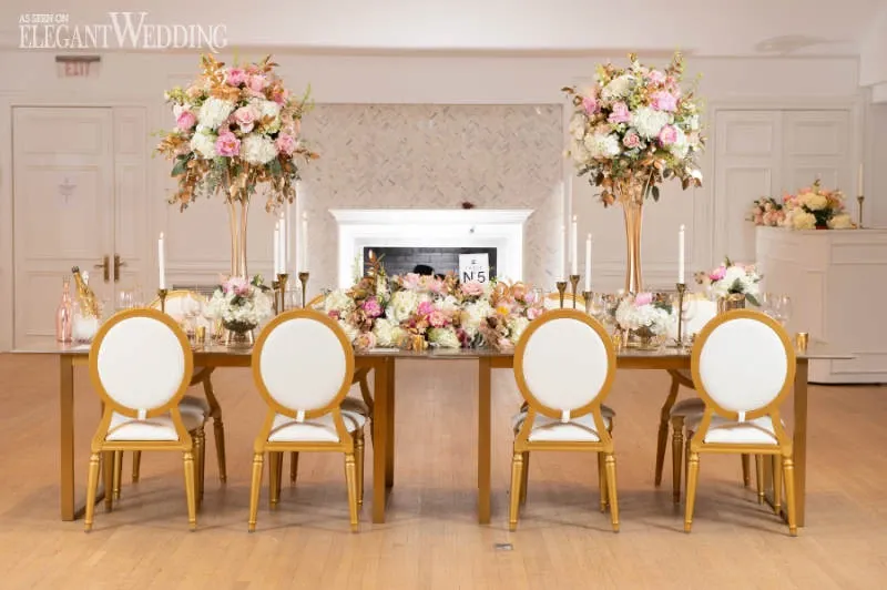 This image showcases an elegantly decorated wedding reception table by La Grâce des Fleurs, categorized under Wedding Florists. The table features a luxurious arrangement with two tall floral centerpieces standing on golden stands, filled with pink, white, and peach flowers that add a touch of romance. Smaller matching floral arrangements are placed strategically along the table, complementing the larger displays. Gold and white chairs surround the table, enhancing the sophisticated aesthetic. Candles and delicate tableware complete the setup, creating a perfect setting for an elegant wedding celebration. The backdrop of the reception area is tastefully subtle, allowing the floral designs to stand out beautifully.