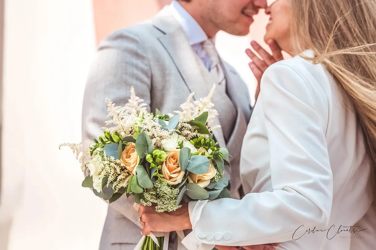 A newlywed couple shares an intimate moment as they lean in for a kiss, with soft smiles on their faces. The bride, dressed in an elegant white suit, gently holds a bouquet of peach roses, white astilbe, and greenery, reflecting the elegance of the occasion. The groom is captured in a stylish light gray suit, adding a touch of sophistication to the scene. This beautiful moment is skillfully captured by Caroline Clouâtre photographe, specializing in wedding photography.