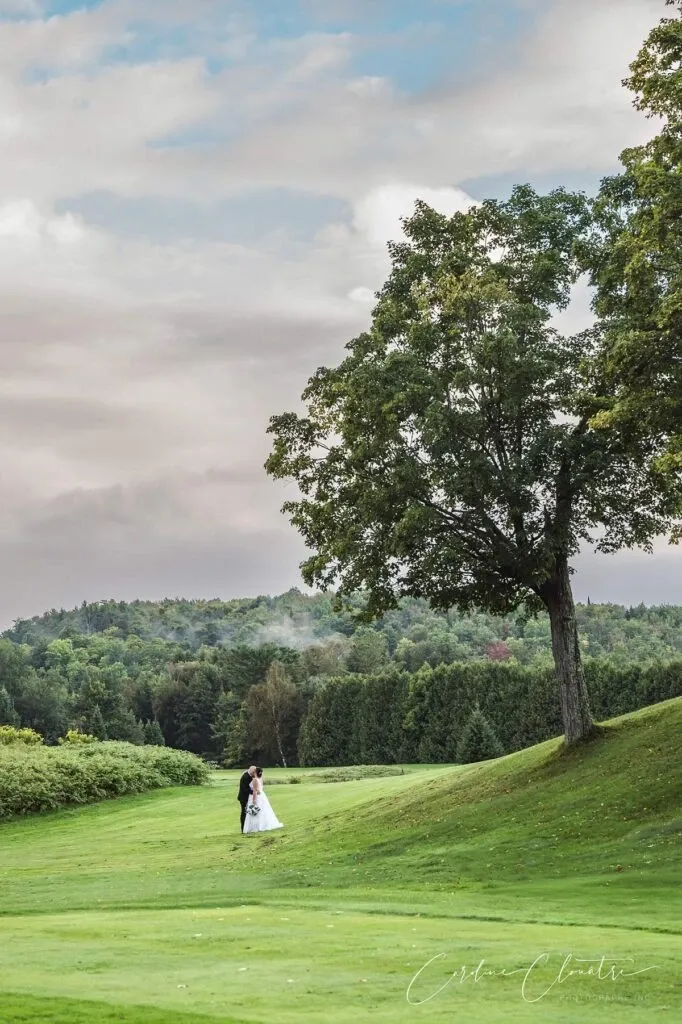 In this serene wedding photograph by Caroline Clouâtre photographe, a bride and groom are seen walking hand in hand across a lush green field. The couple is elegantly dressed, capturing the essence of their special day under a vast, slightly cloudy sky. A large majestic tree adds a picturesque element to the scene, symbolizing growth and union. This image beautifully showcases the art of wedding photography, highlighting the natural beauty and emotional connection of the moment.