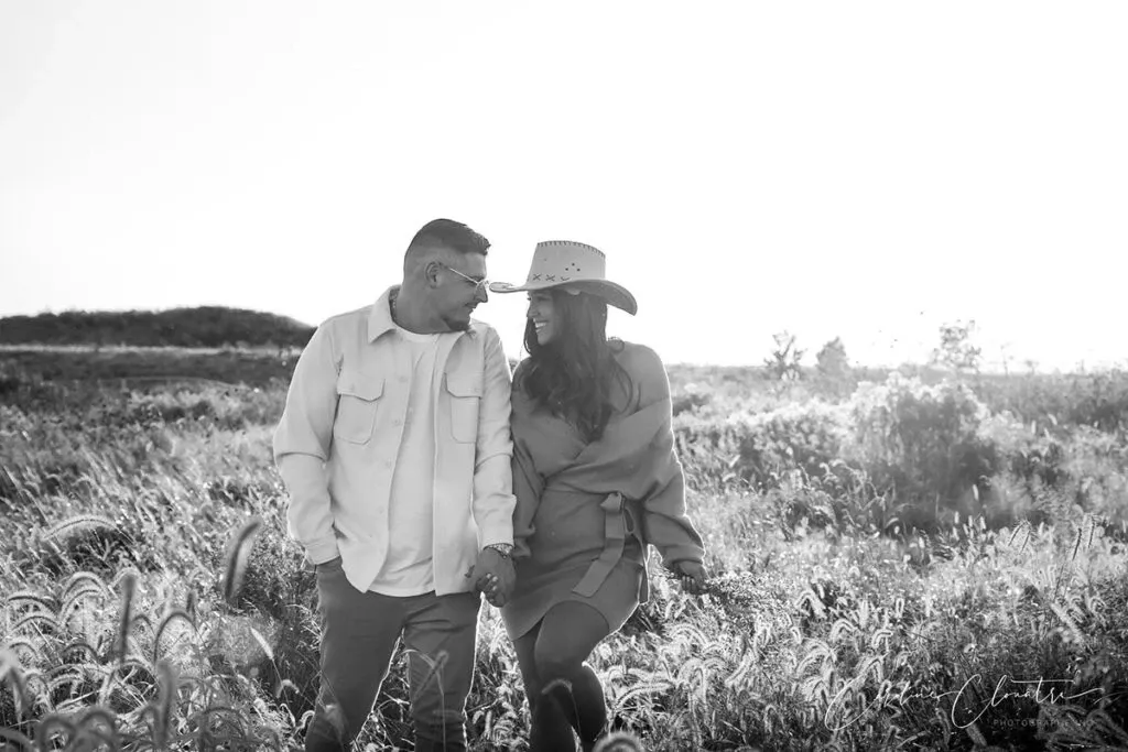 In this black-and-white image by Caroline Clouâtre photographe, specializing in wedding photography, a couple is joyfully walking hand in hand through a sunlit field. The woman is wearing a stylish hat and a cozy outfit, while the man is dressed in a casual shirt and glasses. They share a happy, intimate moment, smiling warmly at each other. The landscape is filled with tall grass and softly blurred sunlight, creating a romantic, timeless atmosphere, perfect for wedding photography.