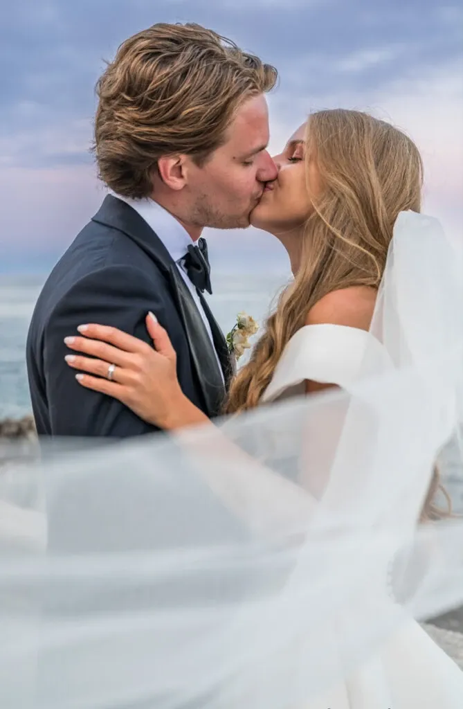 A bride and groom share a romantic kiss by the ocean under a soft, cloudy sky. The bride, dressed in an elegant white off-shoulder gown and a flowing veil, gently holds the groom, who is attired in a classic dark suit and bow tie. Her long, blonde hair cascades gracefully over her shoulders. The veil elegantly drapes over them, adding a dreamy quality to the scene. This intimate wedding moment captures the artistry and style of Caroline Clouâtre photographe, a featured vendor in the Wedding Photography category on Mariage Elegant, Canada.