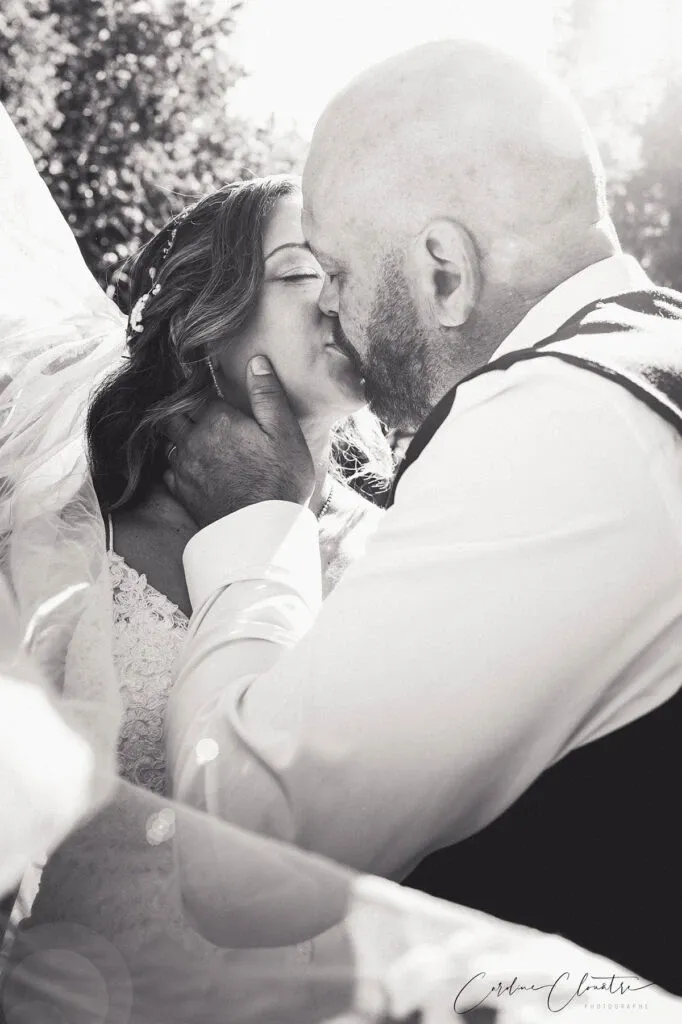 A bride and groom share a tender kiss in an outdoor setting, surrounded by soft sunlight filtering through the trees. The bride is adorned in a lace wedding gown and a delicate veil, while the groom wears a formal vest. The image captures a moment of intimacy and love, emphasizing the couple's connection. This black-and-white photograph highlights the artistic style of Caroline Clouâtre photographe, a wedding photography vendor known for capturing genuine emotions.