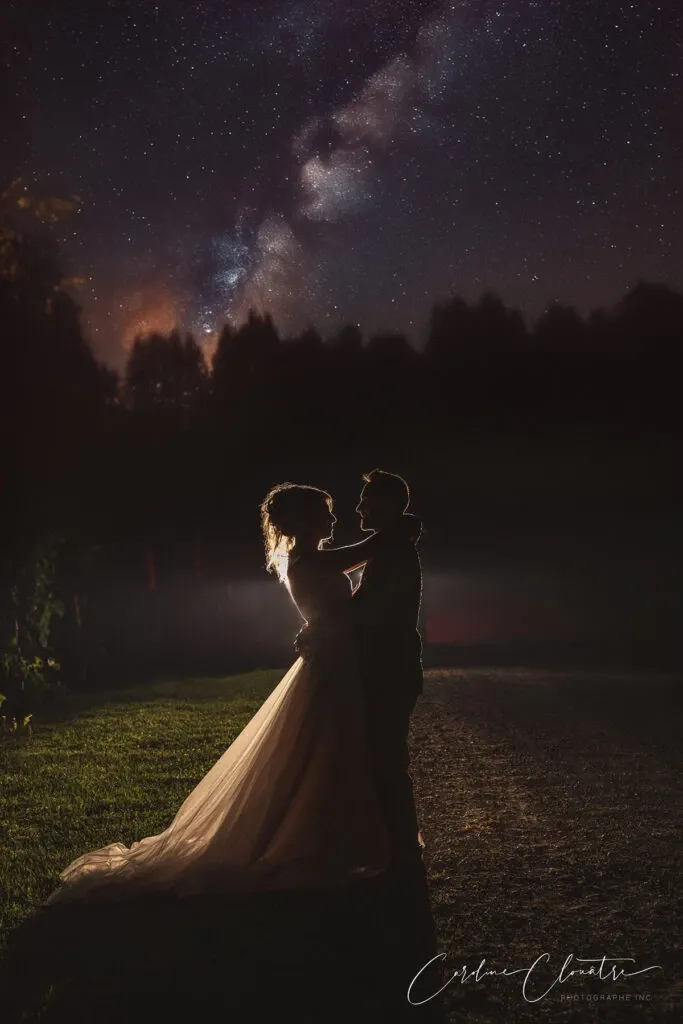 A bride and groom stand together in an open outdoor setting, embracing under a star-lit sky with the Milky Way visible in the background. The silhouettes of the couple are delicately illuminated, highlighting the bride's flowing gown and elegant hairstyle. The romantic scene captures a moment of intimacy and wonder, beautifully photographed by Caroline Clouâtre photographe, a vendor in the Wedding Photography category for Mariage Elegant in Canada.