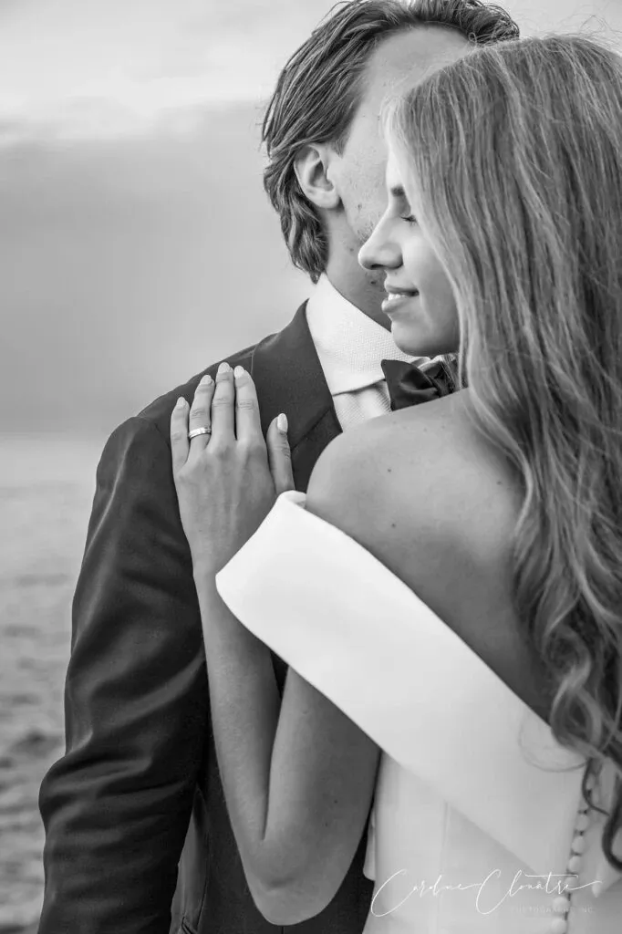 This black and white photograph, captured by Caroline Clouâtre photographe, showcases an intimate moment between a bride and groom, skillfully highlighting their wedding day. The bride, with long, flowing hair, gently places her hand on the groom's shoulder, drawing attention to her elegant engagement ring. Her off-the-shoulder white wedding gown exudes classic elegance. The groom, in a sharp suit and bow tie, stands close, creating a romantic silhouette. The serene backdrop of water adds a timeless and graceful element to this beautiful wedding photography moment by Caroline Clouâtre.