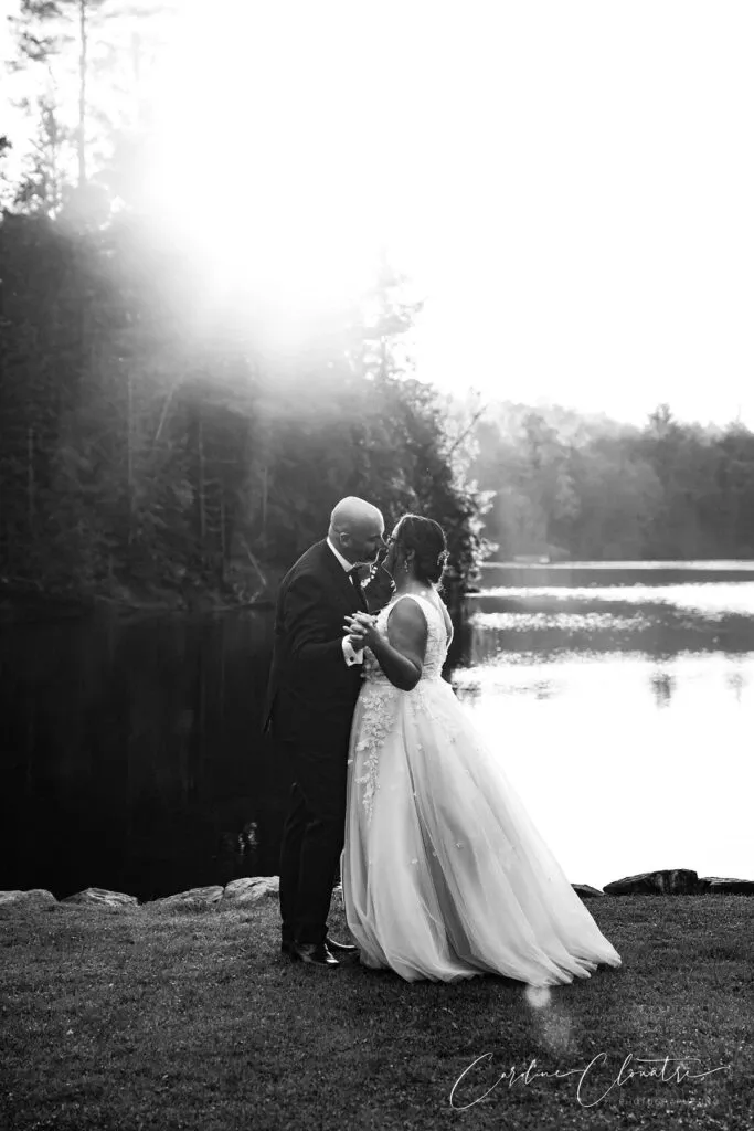 A bride and groom share an intimate moment by a serene lakeside in the soft glow of the setting sun. The bride, dressed in a flowing, ornate wedding gown, gazes lovingly at the groom, who is elegantly attired in a classic suit. The tranquil water and surrounding trees create a picturesque and romantic backdrop, highlighting the beauty of nature. This captivating moment is captured by Caroline Clouâtre photographe, showcasing her expertise in the wedding photography category.