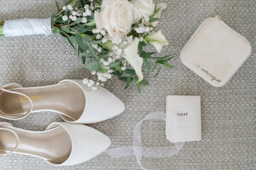 This image, captured by Olivier LG Photographe, a wedding photography expert from Mariage Elegant, Canada, displays a beautifully arranged bridal flat lay. The composition features a bouquet of delicate white roses and greenery, elegantly wrapped with ribbon. Alongside the bouquet are a pair of elegant white bridal heels, perfectly complementing the setup. A personalized clutch and a handkerchief with 'vœux' inscribed on it add a touch of personalized charm. This setup exudes sophistication and is ideal for capturing the essence of a wedding day.