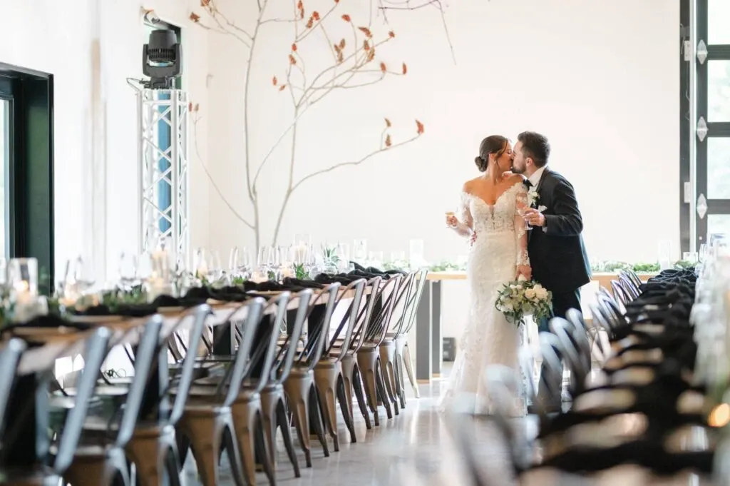 In this elegant wedding photograph by Olivier LG Photographe, categorized under Wedding Photography, a newlywed couple is captured sharing a tender kiss. The bride is wearing an exquisite lace gown, holding a bouquet of white flowers, while the groom is dressed in a classic dark suit with a boutonnière. They are standing at the end of a beautifully set table adorned with greenery and candles, creating a romantic ambiance. The room features modern decor with a minimalist aesthetic, enhanced by natural light streaming through large windows. This image perfectly encapsulates the sophistication and joy of a wedding celebration in Canada.
