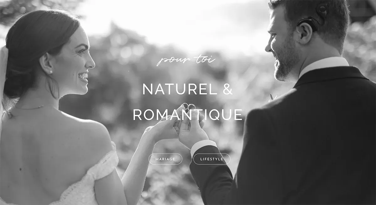 The black and white image shows a bride and groom facing each other and holding hands, surrounded by soft, diffused natural lighting that creates a romantic atmosphere. The bride, wearing an elegant wedding dress with off-the-shoulder sleeves, smiles at the groom, who is dressed in a classic suit. In the center of the image, the text reads 'pour toi,' 'NATUREL & ROMANTIQUE,' highlighting the theme of the photo. The words 'Mariage' and 'Lifestyle' are also visible, indicating the wedding context. This image showcases the artistic and emotive style of Olivier LG Photographe, a vendor in the Wedding Photography category for Mariage Elegant, Canada.