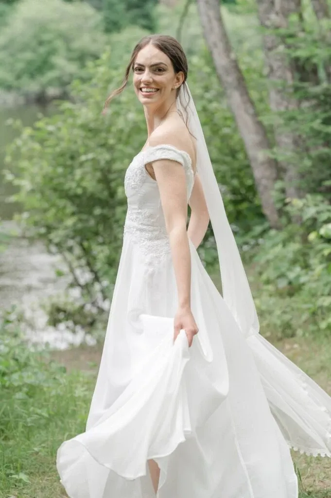 In this image by Olivier LG Photographe, a bride is elegantly captured in a picturesque outdoor setting, showcasing the artistry of wedding photography. She is wearing a beautiful white gown with delicate lace details and an off-the-shoulder design. Her veil cascades down her back as she joyfully turns, highlighting the flowy skirt of her dress. The background features lush greenery and a serene body of water, adding a natural and tranquil touch to the scene. This image perfectly exemplifies the expertise in capturing candid, joyful wedding moments, making it an ideal representation of the wedding photography category for Mariage Elegant in Canada.