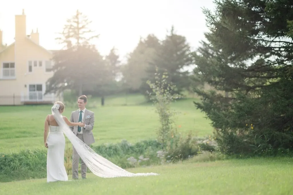 A bride and groom stand together in a lush green field, looking into each other's eyes. The bride is wearing an elegant white gown with a long, flowing veil, while the groom is dressed in a stylish suit. In the background, there is a large house, partially obscured by tall trees, creating a serene and romantic atmosphere. This image captures a tender wedding moment, showcasing the work of Olivier LG Photographe, a vendor specialized in wedding photography, listed on Mariage Elegant in Canada.
