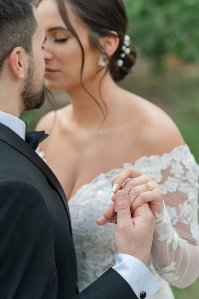 A close-up image of a bride and groom embracing tenderly, taken by Olivier LG Photographe, a wedding photography vendor. The bride is wearing an elegant, off-the-shoulder lace wedding gown with delicate floral designs and pearl adornments in her updo. The groom is dressed in a classic black tuxedo with a white shirt and black bow tie. They are holding hands gently, showcasing the bride's wedding ring, with the lush greenery serving as a soft-focused backdrop, creating a romantic atmosphere. This image beautifully captures the intimate and loving moment typical of a wedding day.