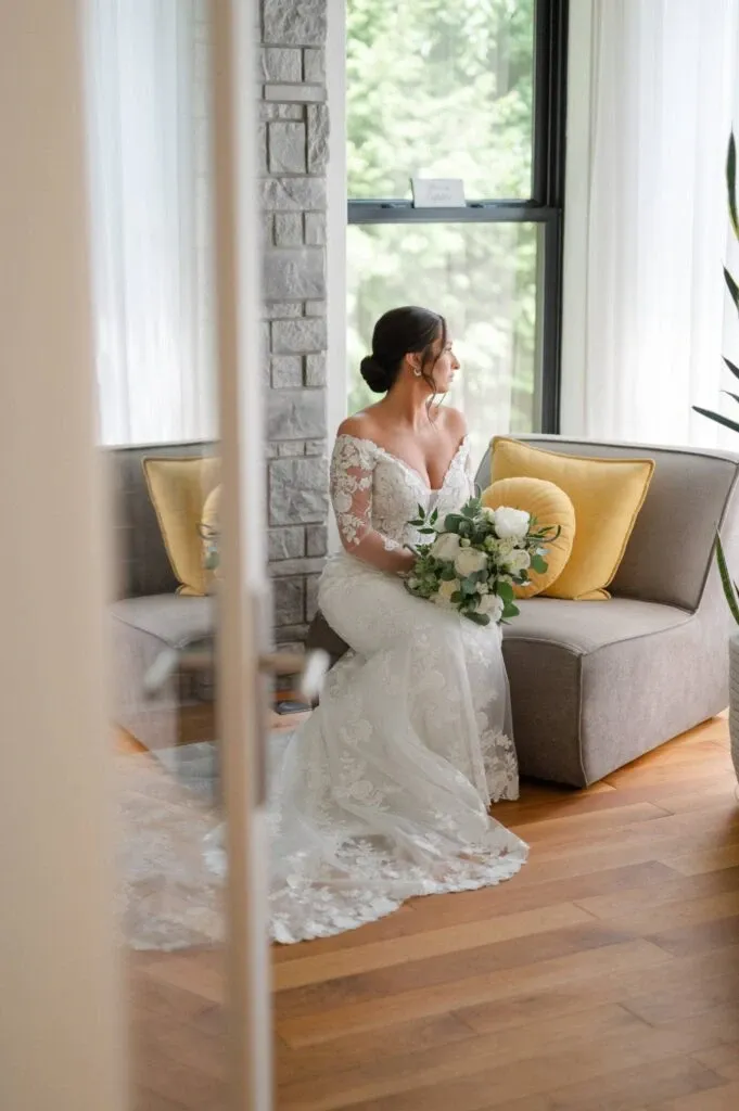 A bride in an elegant lace wedding gown sits gracefully on a modern gray sofa with yellow cushions. She holds a lush bouquet of white and green flowers and gazes thoughtfully out a large window, allowing natural light to illuminate the scene. The setting features wooden floors and a stone-textured wall, creating a sophisticated and intimate atmosphere. This image, captured by Olivier LG Photographe, showcases the artistry of wedding photography in creating timeless and romantic moments.