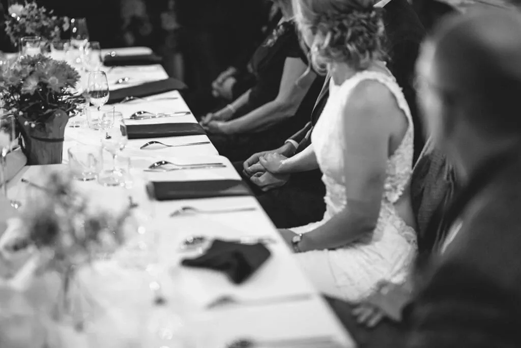 This black and white image by Olivier LG Photographe, featured in the Wedding Photography category on Mariage Elegant, Canada, captures a serene moment at a wedding reception. A bride, wearing an elegantly detailed lace gown, is seated at a long table adorned with neatly arranged cutlery, napkins, and floral centerpieces. The soft focus showcases guests engaged in quiet conversation, creating an intimate and sophisticated atmosphere typical of a refined wedding celebration.
