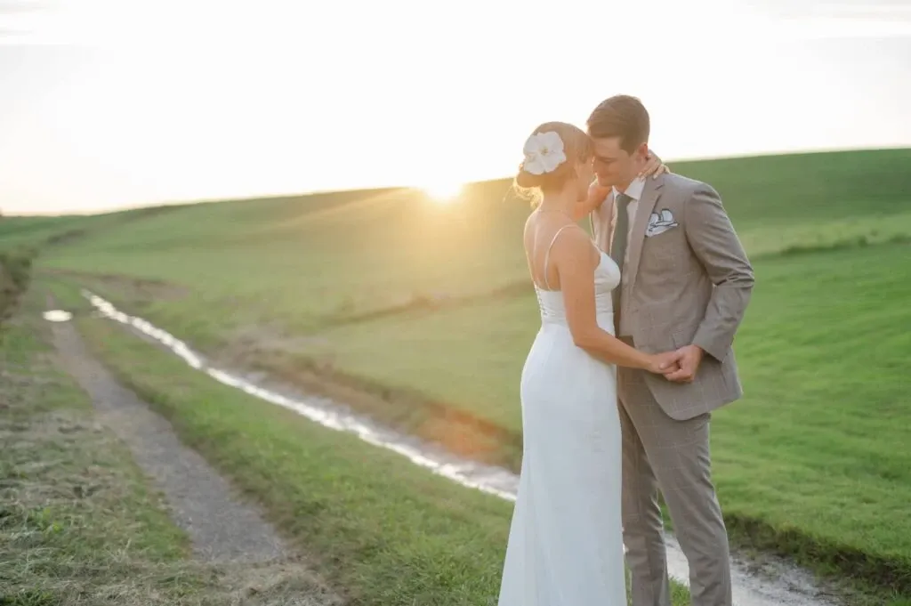 A bride and groom stand in a serene outdoor setting at sunset, captured by Olivier LG Photographe, a vendor in the Wedding Photography category. The bride is wearing an elegant white gown with a large white floral hairpiece, while the groom is in a light gray suit, complete with a checkered pocket square. They share an intimate moment, holding hands along a grassy path next to a gentle stream, with the sun setting in the background, creating a warm glow over the picturesque landscape.