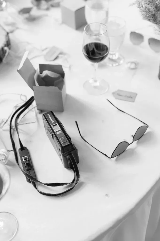The image shows a tabletop scene at a wedding, featuring a vintage camera with a strap, positioned next to a pair of sunglasses. A partially opened small gift box is also visible, suggesting it's part of a wedding favor. Nearby, there are a glass of red wine and several place cards, enhancing the setting's elegance. This photograph, captured by Olivier LG Photographe, falls under the wedding photography category for Mariage Elegant in Canada.