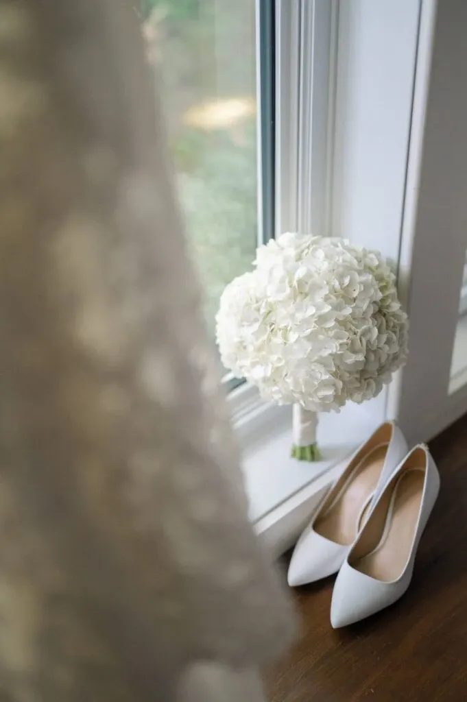This image showcases a wedding scene captured by Olivier LG Photographe, a vendor in the Wedding Photography category. It features elegant white high-heeled shoes placed on a wooden floor next to a delicate bouquet of white hydrangeas. The soft, blurred foreground suggests bridal attire, adding a romantic ambiance that complements the natural light streaming through a nearby window. This setup highlights the attention to detail and style characteristic of wedding preparations.