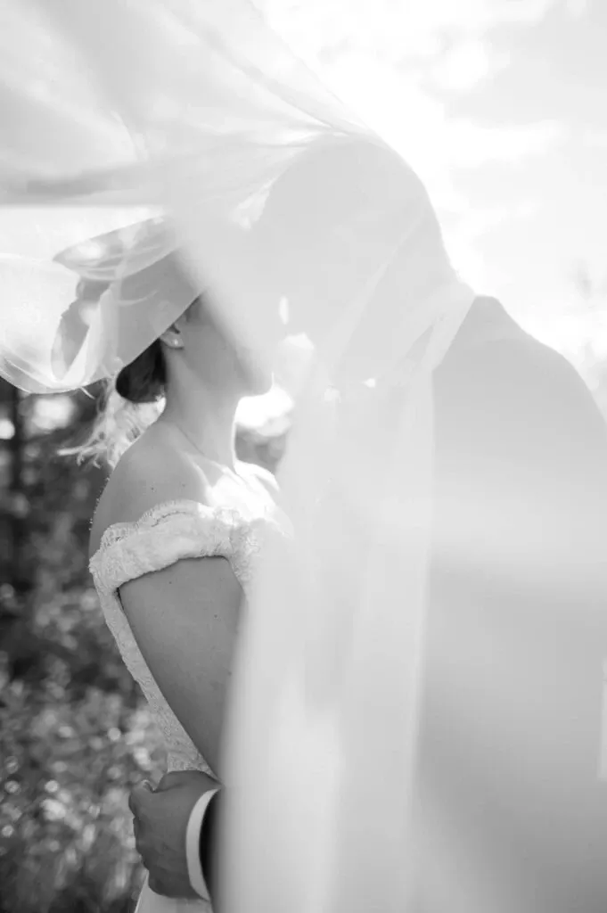 This elegant black-and-white image captured by Olivier LG Photographe, a vendor in the Wedding Photography category, showcases a romantic moment between a bride and groom beneath a flowing veil. The bride is wearing a delicate off-the-shoulder wedding gown, while the groom is in a classic suit, both softly illuminated by natural sunlight. This intimate scene embodies the essence of love on a wedding day, beautifully highlighting the artistry of skilled wedding photography.