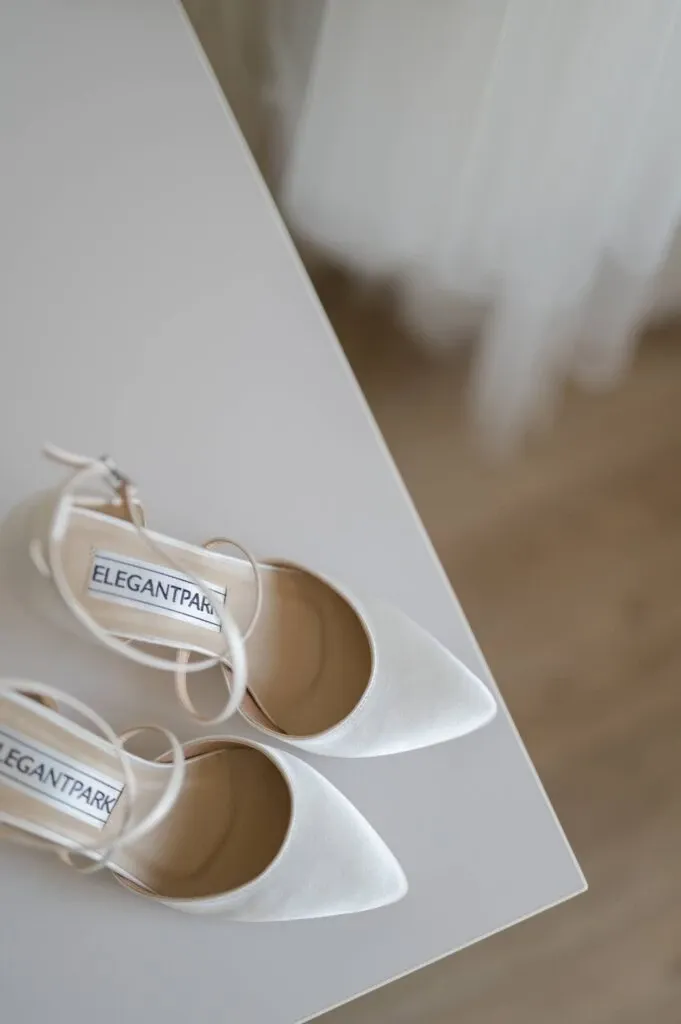 In this image, a pair of elegant white bridal shoes is neatly arranged on a light-colored surface, labeled with the brand 'Elegantpark' on the insole. The shoes have a pointed toe and delicate ankle straps, ideal for a sophisticated wedding look. In the blurred background, a glimpse of a wedding dress adds to the bridal theme. This photograph, taken by Olivier LG Photographe, features in the wedding photography category on Mariage Elegant, Canada, showcasing attention to detail and style.