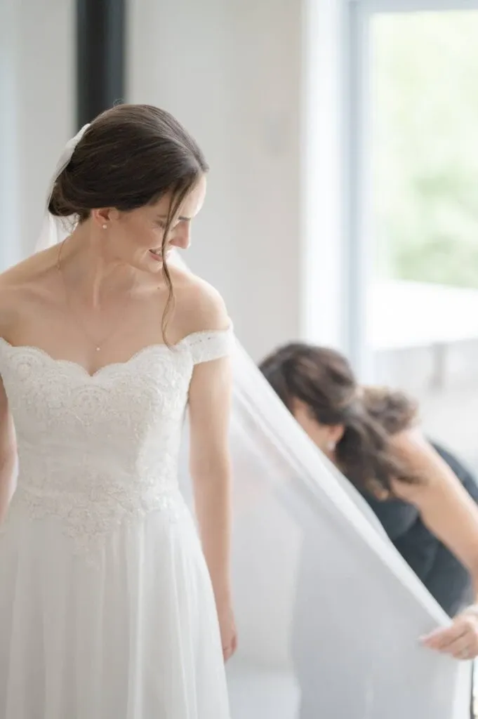 A bride, captured by Olivier LG Photographe, a vendor specializing in wedding photography, stands gracefully in an elegant, off-the-shoulder lace wedding dress. Her hair is styled in a neat updo with a veil cascading down her back. A woman, possibly a bridesmaid or stylist, assists by adjusting the veil, adding to the serene, intimate preparation moment. Soft natural light fills the space, suggesting a calm and beautifully lit environment, perfect for wedding preparations in Canada.