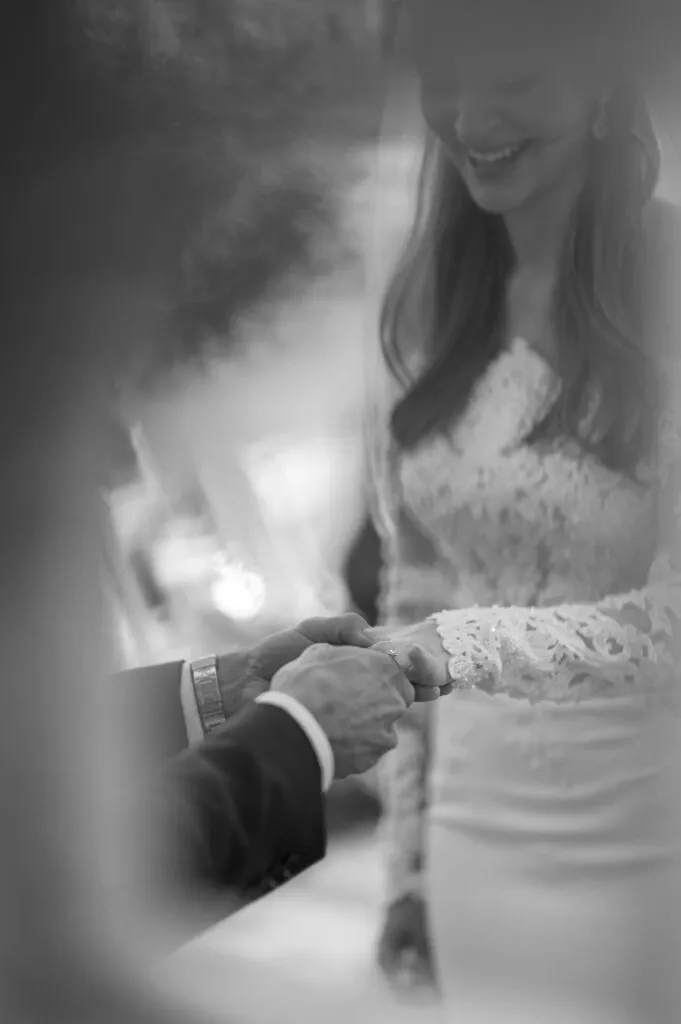 In this black and white wedding photograph by Olivier LG Photographe, a bride and groom are showcased exchanging vows. The image captures an intimate moment as the couple holds hands, with the bride wearing a lace-sleeved gown and smiling gently. The groom's hand is adorned with a watch, hinting at a timeless elegance, while the soft focus around the edges of the image creates a dreamy, romantic atmosphere. This photo beautifully encapsulates the essence of wedding photography and demonstrates the artistry of capturing emotions on a couple's special day.