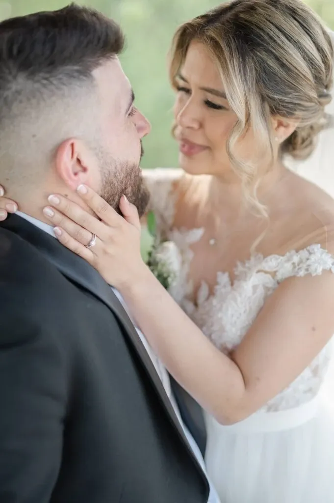 A joyous bride and groom captured in an intimate moment on their wedding day. The bride, wearing a delicate lace wedding dress, gently holds the groom's face with both hands, showcasing her elegant engagement ring. Her hair is styled in soft curls, and she wears a graceful veil. The groom is dressed in a classic black suit, gazing back at her with affection. This image by Olivier LG Photographe perfectly encapsulates the love and connection between the couple, emphasizing the expertise in wedding photography.