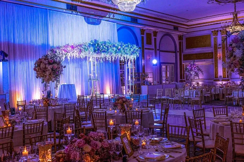 This image showcases an elegantly decorated wedding reception venue captured by Guy Chan Chive Photography, a vendor in the Wedding Photography category. The setting features a spacious ballroom with opulent chandeliers hanging from the ceiling, casting a warm glow over the room. A beautifully arranged floral arch, adorned with cascading white and lavender flowers, serves as a focal point, set against a backdrop of flowing white drapes illuminated with soft purple lighting. The tables are meticulously set with glassware and floral centerpieces, enhancing the romantic and luxurious ambiance of the event. This reflects the sophisticated style associated with Mariage Elegant in Canada.