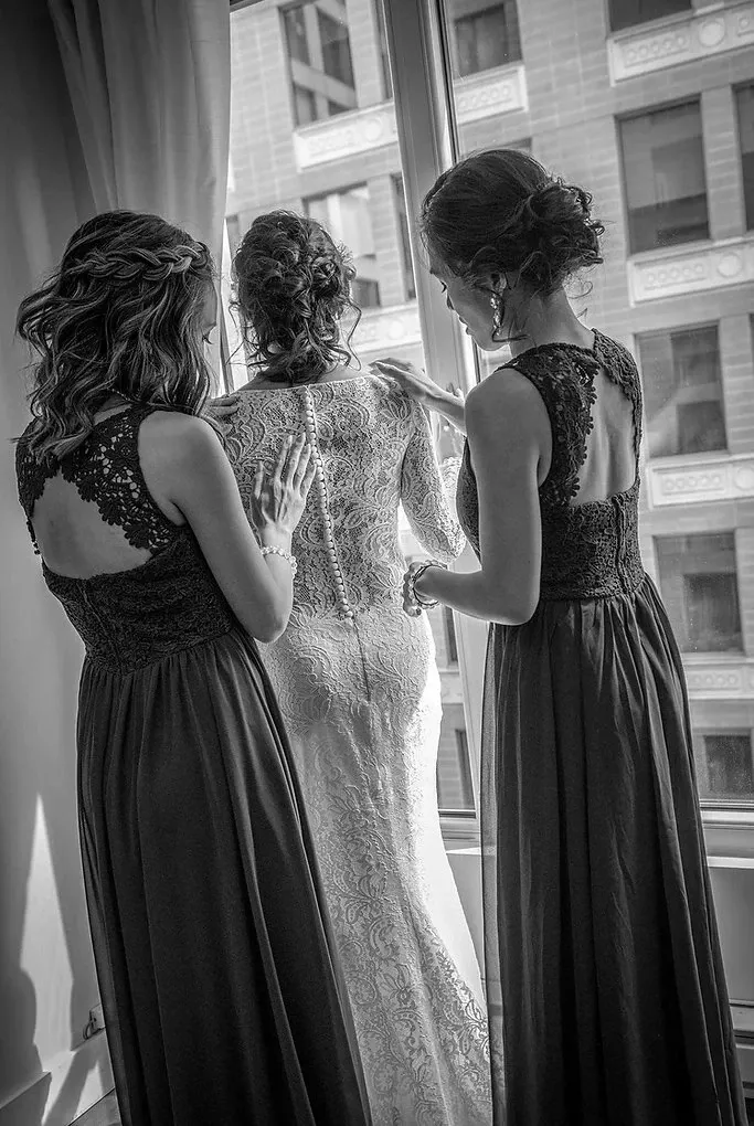 In this black-and-white image by Guy Chan Chive Photography, part of the wedding photography category on Mariage Elegant, Canada, a bride in an intricately detailed lace wedding gown stands by a bright window. Two bridesmaids, also dressed in elegant lace gowns, assist her with the final adjustments of her dress. The scene captures a tender moment of preparation in a high-rise building, highlighting the elegant and intimate atmosphere typical of a sophisticated wedding setting.