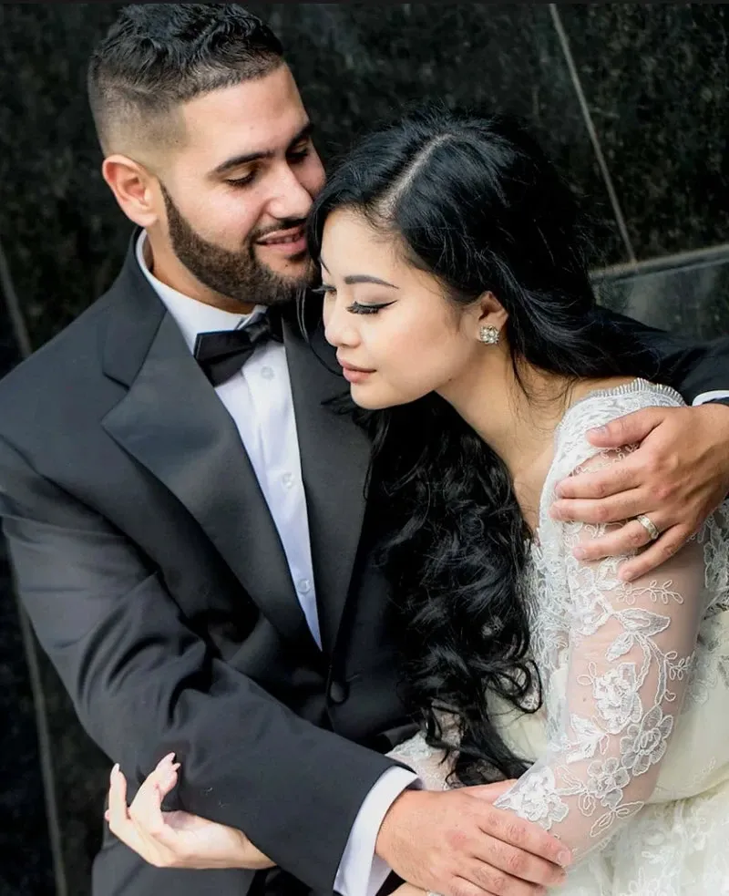 A couple lovingly embraces in an elegant setting, showcasing the fine artistry of Guy Chan Chive Photography, a wedding photography vendor on Mariage Elegant, Canada. The groom, wearing a classic black tuxedo complemented by a bow tie, gently holds the bride in his arms. The bride is adorned in a lace wedding gown with intricate detailing, her long dark hair cascading over her shoulder. This intimate moment captures the essence of romance and elegance, perfect for a wedding day celebration.