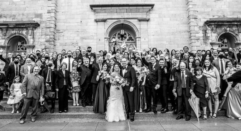 A large, joyful wedding party is gathered on the steps of a grand stone building. The couple, dressed in a classic wedding gown and a sharp suit, stands smiling at the center, surrounded by friends and family. The group displays a variety of expressions, from cheerful smiles to joyous laughter. The historical architecture of the building adds an elegant backdrop to the celebratory scene. This image showcases the wedding photography skills of Guy Chan Chive Photography, capturing a memorable and festive moment in a wedding context for Mariage Elegant in Canada.