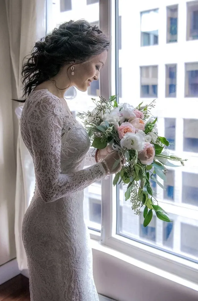 A bride in an elegant lace wedding dress stands by a large window, holding a bouquet of pastel pink and white flowers with lush greenery. The natural light highlights her intricate hairstyle and delicate earrings. This image, captured by Guy Chan Chive Photography, beautifully showcases the artistry of wedding photography, highlighting the intimate and serene moments of a bride on her wedding day. This example of wedding photography is ideal for those seeking timeless and sophisticated imagery for their special event.