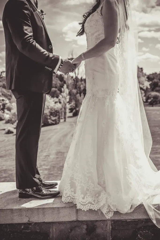A bride and groom stand outdoors, holding hands and facing each other, captured in an elegant black-and-white photograph by Guy Chan Chive Photography, a vendor in the wedding photography category. The bride wears a lace-embellished gown with a long veil that cascades down her back. The groom is dressed in a classic suit. They are positioned on a stone ledge with a scenic background of a landscaped garden and a bright sky, adding a romantic ambiance to the moment. This image is ideal for showcasing the timeless beauty of wedding photography offered by Guy Chan Chive Photography at Mariage Elegant in Canada.