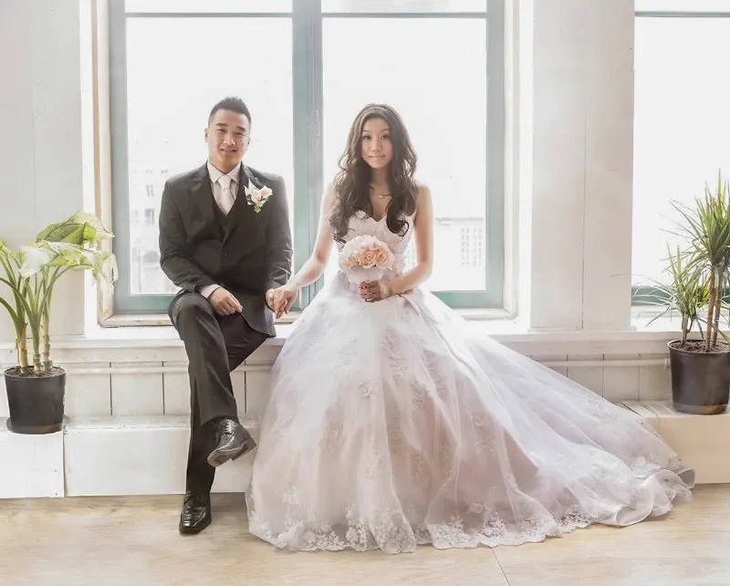 This image showcases a newlywed couple sitting beside each other in a bright, sunlit room, captured by Guy Chan Chive Photography for the wedding photography category. The bride, in an elegant white lace gown, holds a bouquet of pale pink roses, while the groom is dressed in a sleek dark suit with a matching boutonniere. They are framed by large windows and flanked by potted plants, adding a touch of natural greenery to the serene setting. This photograph highlights the couple's joyous moment, embodying the essence of a sophisticated wedding celebration.