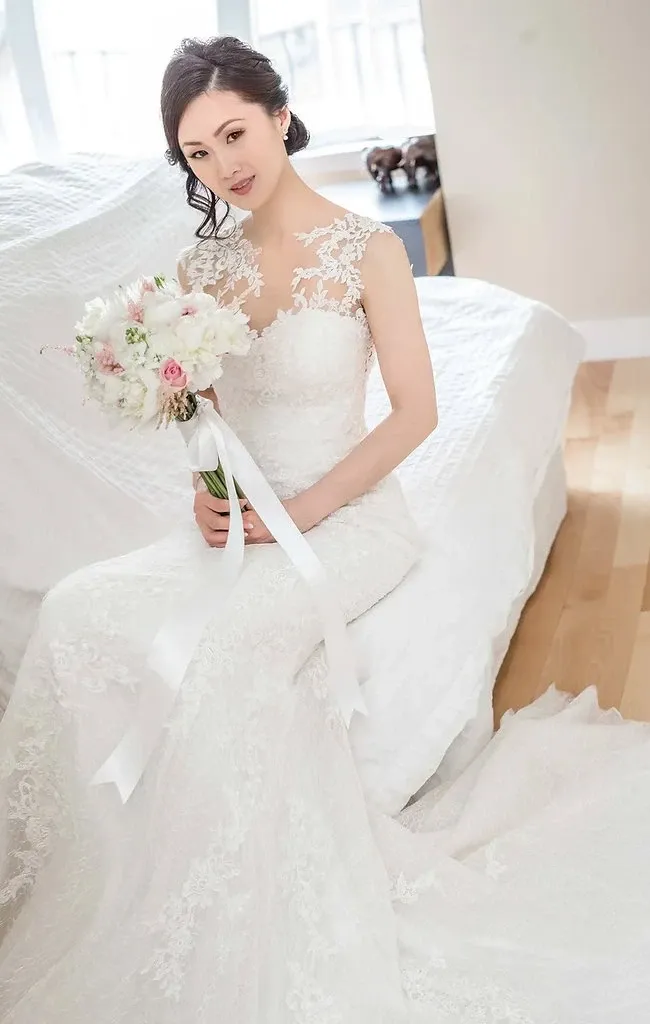 A bride sits gracefully on the edge of a bed, wearing an elegant lace wedding gown. The room is softly lit, accentuating the intricate floral lace details of her dress. She holds a delicate bouquet of white and pink flowers, tied with a long white ribbon. Her hair is styled in loose waves, and she gazes softly towards the camera, embodying a serene bridal elegance. This image captures the artistry of Guy Chan Chive Photography, showcasing their expertise in wedding photography for Mariage Elegant, Canada.