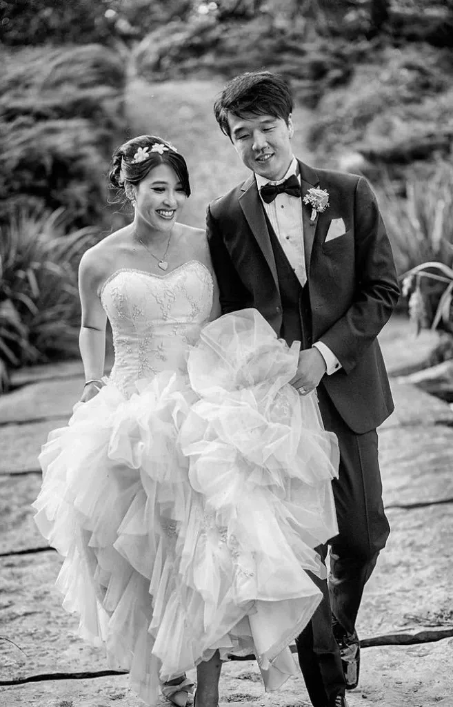 A joyful bride and groom walk hand-in-hand down a rustic stone path surrounded by lush greenery. The bride is wearing a strapless, tiered wedding gown and holds it up slightly to ease her steps. Her hair is styled elegantly with floral accents matching her delicate necklace. The groom, dressed in a classic black tuxedo with a bow tie and boutonniere, looks contently at his bride. This image, captured by Guy Chan Chive Photography, perfectly embodies the romance and sophistication of wedding photography, suitable for a memorable Canadian wedding.