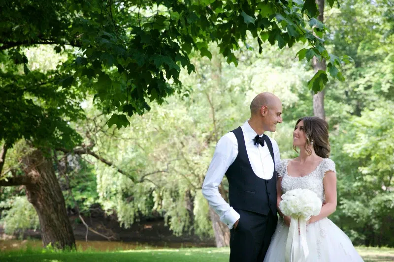 A bride and groom stand in a lush, green outdoor setting beneath a large tree, exchanging a joyful glance. The bride is wearing a white, lace wedding dress and holding a bouquet of white flowers, while the groom is in a black tuxedo with a bow tie. The background features trees and a gently flowing river, creating a serene and romantic atmosphere. This image showcases the wedding photography services of Studio Omega Photo under the wedding category on Mariage Elegant, Canada.
