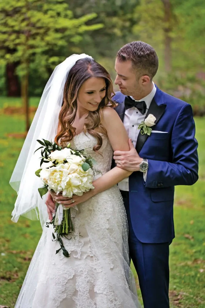 A bride and groom are standing together in a lush, green garden, the bride wearing an elegant lace wedding dress and a flowing veil, holding a bouquet of white flowers accented with greenery. The groom is dressed in a sharp blue suit with a black bow tie and a white boutonniere, gently embracing the bride as they both smile warmly. This romantic scene captures a moment of joy and love, perfectly photographed by Studio Omega Photo, a featured vendor in the Wedding Photography category on Mariage Elegant, Canada.