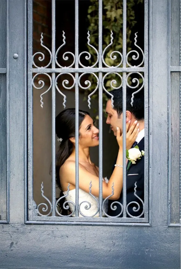 In a captivating photograph by Studio Omega Photo, a vendor specializing in wedding photography under Mariage Elegant in Canada, a bride and groom share a tender moment framed by intricate wrought iron gates. The bride lovingly cups the groom's face, both smiling and gazing into each other's eyes. The bride is adorned in a strapless white wedding gown, and her dark hair is elegantly styled. The groom is dressed in a formal black suit and white shirt, with a boutonniere pinned to his lapel, enhancing the romantic ambiance of their wedding day.