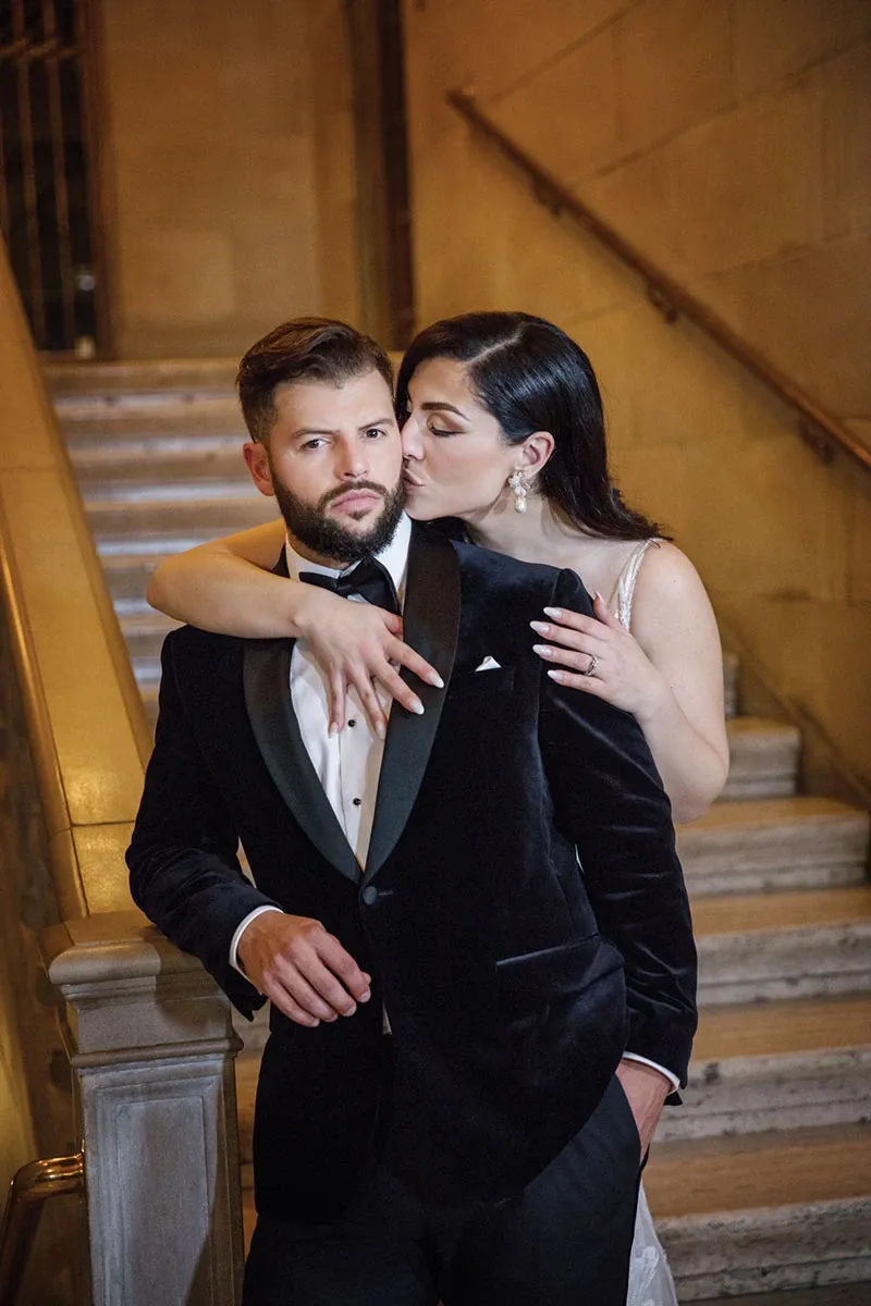In this elegant wedding photography image by Studio Omega Photo, a groom and bride are posed on a grand staircase. The groom is dressed in a classic black velvet tuxedo with a bow tie, exuding sophistication and style. The bride embraces him from behind, wearing a shimmering gown, her hair styled elegantly. They are positioned against the backdrop of ornate stone railings, capturing a timeless moment. This image exemplifies the exquisite photography skills offered by Studio Omega Photo, specializing in wedding photography for Mariage Elegant, Canada.