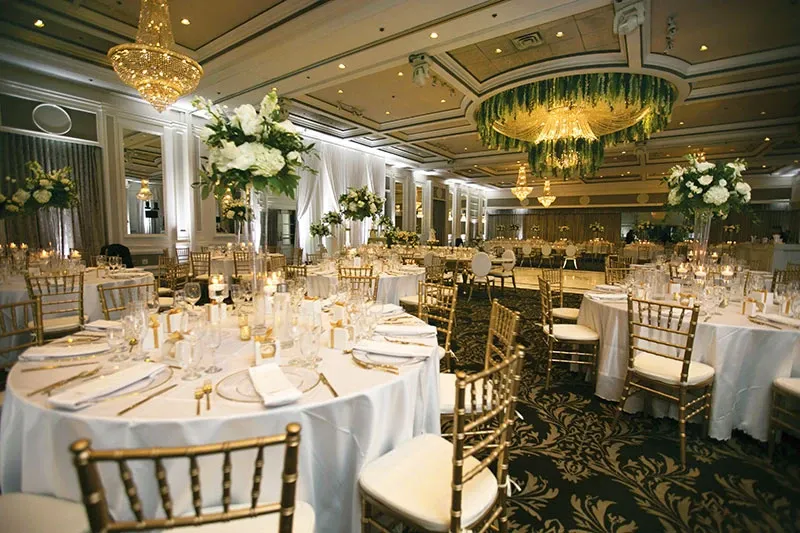 The image showcases an elegantly decorated wedding reception hall, captured by Studio Omega Photo, highlighting their expertise in wedding photography. The venue features beautifully arranged round tables with white tablecloths, gold chiavari chairs, and centerpieces of lush white and green floral arrangements. A stunning chandelier with cascading greenery serves as the focal point of the room, adding a touch of sophistication. Soft ambient lighting and numerous candles create a warm and inviting atmosphere, ideal for a wedding celebration.