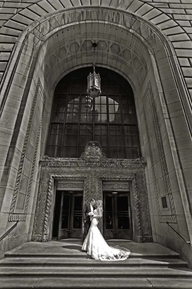 This image captures a newlywed couple in an elegant pose in front of a grand, intricately carved stone entrance, part of an impressive architectural structure. The bride, in a flowing, detailed wedding gown, and the groom, in a classic tuxedo, gaze lovingly at each other, adding a romantic touch to the scene. The photograph, taken by Studio Omega Photo, showcases their exceptional work in the wedding photography category. The couple is positioned under a large arched window with ornamental patterns, enhancing the majestic ambiance perfect for a memorable wedding album.
